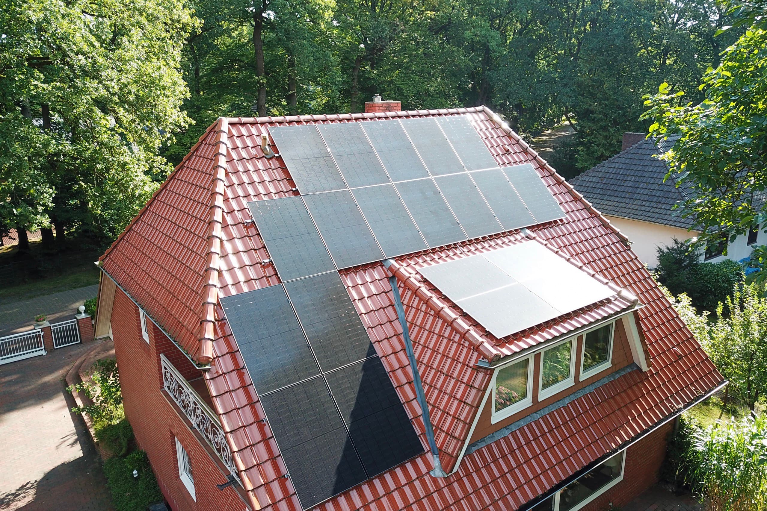 Drohnenaufnahme eines Klinkerhauses mit Walmdach und Photovoltaikmodulen auf zwei Dachseiten, umgeben von Bäumen